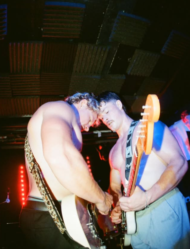 Two muscular men playing electric guitars on stage under bright lights.