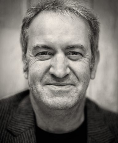 Smiling man with short grey hair, wearing a dark striped blazer against a neutral background.