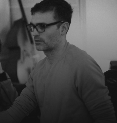 A man in glasses, focused on a task, with a blurred background of musical instruments.
