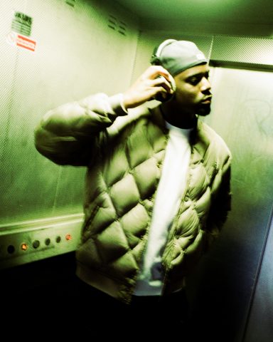 A man wearing a beanie and puffer jacket stands in a dimly lit elevator.